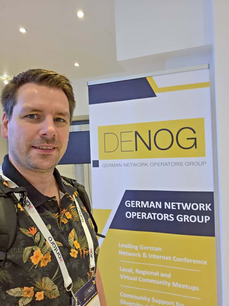 denog17_stefan xCrew-Mitglied Stefan vor einem Roll Up des DENOG e.V.
